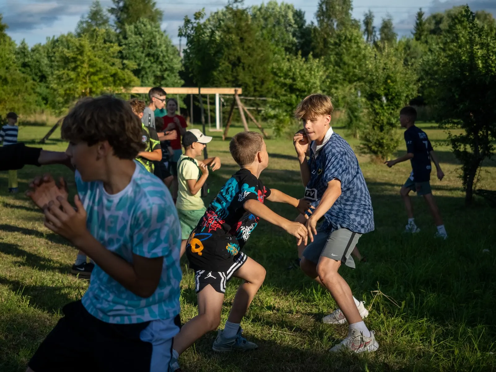 Vaikai žaidžia stovyklos pievoje vakaro saulėje
